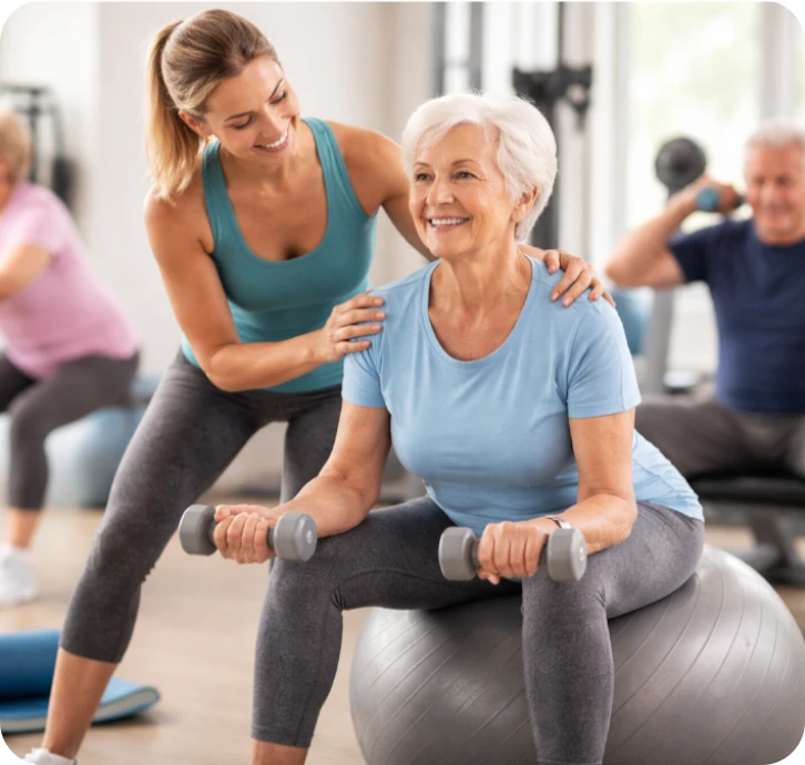 Trainerin unterstützt eine Seniorin bei Übungen mit Kurzhanteln auf einem Gymnastikball